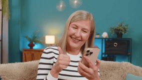 Caucasian mature woman smiles, engages with social media photos videos on smartphone while sitting at home. Grandmother gives thumbs up gesture expressing approval and happiness on sofa in living room - Powered by Shutterstock - Get 15% off with code: PIKWIZARD15