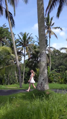 Side view of woman in white dress walking along tropical palm-lined road in Bali, Indonesia. Peaceful island setting with lush greenery, palm trees, and serene atmosphere.