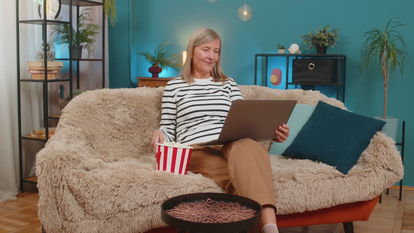 Caucasian mature woman places a bucket of popcorn on table and sets up laptop to watch favorite series film content at home. Grandmother relaxed satisfied, preparing for enjoyable movie night on sofa