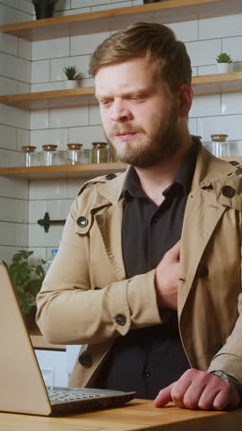 Distressed man clutching his chest in discomfort while sitting in front of a laptop, possibly experiencing a panic attack at home.,vertical video