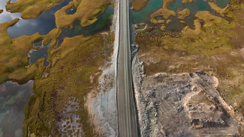 An aerial view of a straight road disappearing into a band of low-lying clouds, surrounded by a rugged, barren landscape with scattered pools of water
