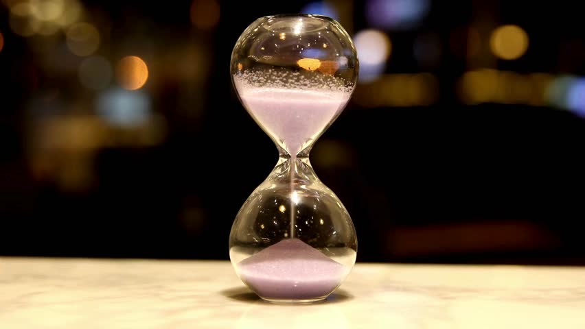 hourglass on table, sand flowing through the bulb. hourglass made of glass in the desk with out-of-focus background. close up of a transparent hourglass with flowing sand on blur background.