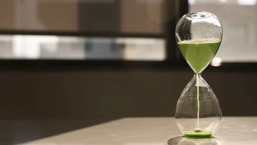 hourglass on table, sand flowing through the bulb. hourglass made of glass in the desk with out-of-focus background. close up of a transparent hourglass with flowing sand on blur background.