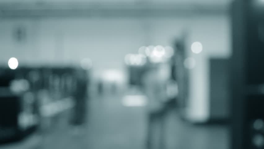 People walking in building. Building with walls, floor, ceiling illuminated lamps. Silhouettes of people indoors. Many people walking. Men and women in tall hall. Exhibition center. Blurred background