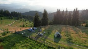 Aerial view of picturesque campsite on grassy hill, surrounded by evergreen trees and overlooking mountain vista. Distant mountains, tourist tents, cars, camping gear under golden evening light. - Powered by Shutterstock - Get 15% off with code: PIKWIZARD15