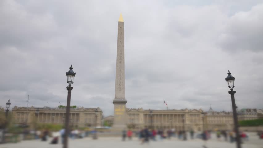 People stroll in the Place de la Concorde with views of the Obelisque de Louxor and Hotel de la Marine in the background on a cloudy spring day in 1st arrondissement. Out of focus. Paris, France.
