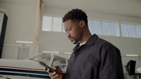 Black male technician in uniform standing in aviation hangar, looking around and checking aircraft-related information on smartphone during workday - Powered by Shutterstock - Get 15% off with code: PIKWIZARD15