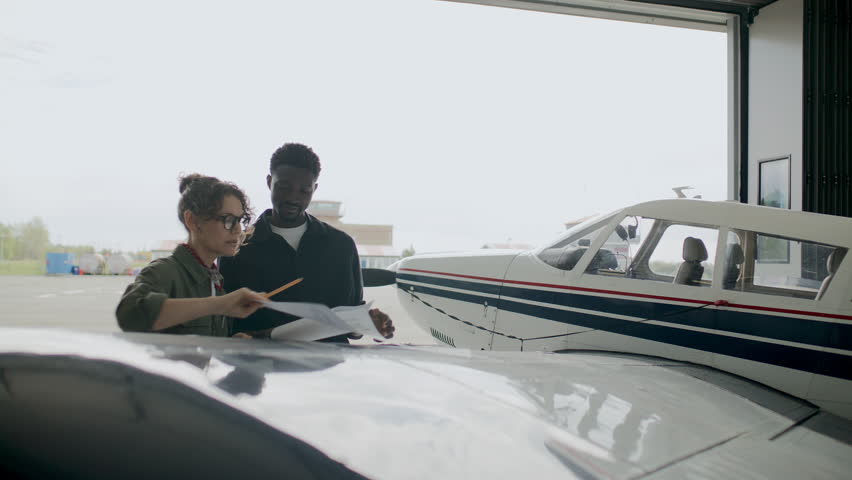 Caucasian female technician and her Black male coworker standing by airplane in open hangar, examining paper plans of aircraft and discussing maintenance details