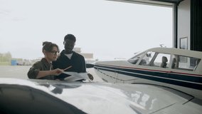 Caucasian female technician and her Black male coworker standing by airplane in open hangar, examining paper plans of aircraft and discussing maintenance details - Powered by Shutterstock - Get 15% off with code: PIKWIZARD15