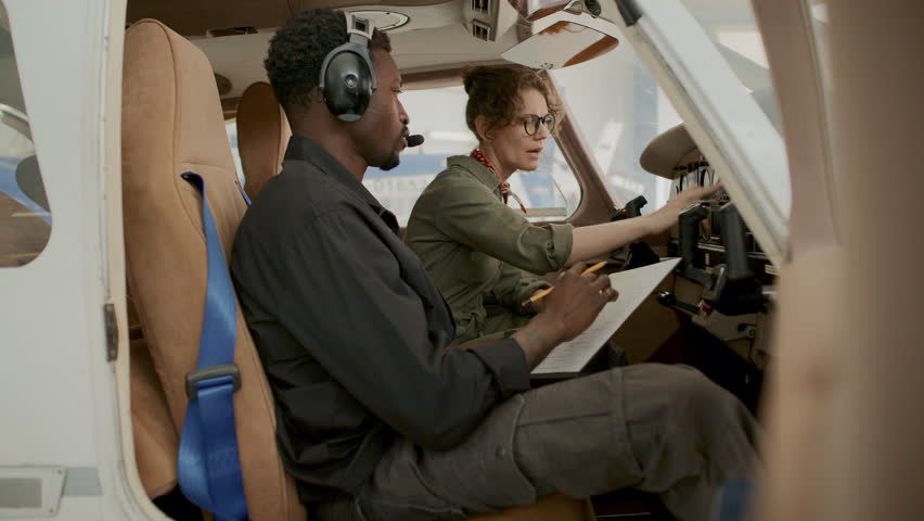 Caucasian female pilot pointing at cockpit controls and explaining instrument functions to Black male technician taking notes on clipboard during preflight aircraft check