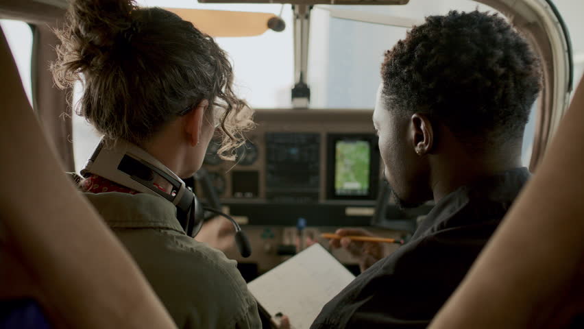 Caucasian female pilot pointing at navigation screen and talking to African American male colleague taking notes during flight training session inside aircraft cockpit