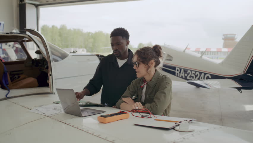 Black male mechanic and Caucasian female technician discussing aircraft repair plan on laptop and paper, standing in hangar by plane wing with multimeter and tools on it