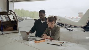 Black male mechanic and Caucasian female technician discussing aircraft repair plan on laptop and paper, standing in hangar by plane wing with multimeter and tools on it - Powered by Shutterstock - Get 15% off with code: PIKWIZARD15