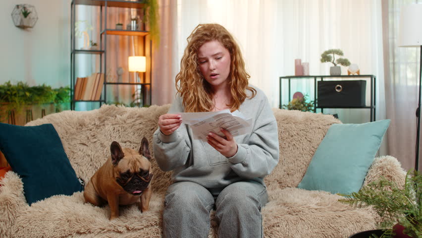 Young Caucasian redhead woman checks bills with serious look while on home sofa, showing signs of stress. Pug dog lies next to her calmly, sensing emotional discomfort and offering quiet presence.