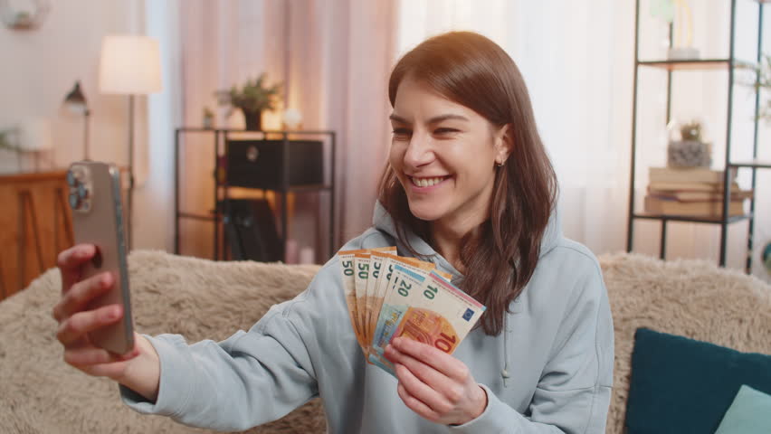 Smiling young woman blogger influencer recording story on smartphone sitting on sofa couch showing money euro banknotes, talking about financial success and easy ways to earn. Girl in casual at home.