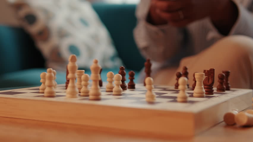 A person moves a chess piece on a wooden chessboard. The focus is on the board and pieces, with the player slightly out of focus.