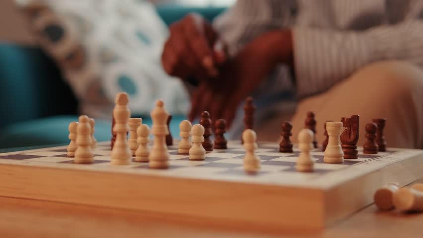 A person moves a chess piece on a wooden chessboard. The focus is on the board and pieces, with the player slightly out of focus.