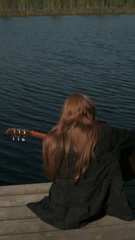 Play Music, classic guitar outdoors, nature lake landscape, travel. Girl with red hair plays guitar. Cinematic 4k vertical