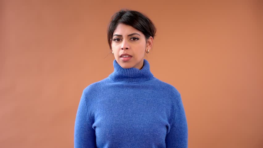 A woman in a blue turtleneck sweater listens intently while tilting her head against brown background during a creative studio session focused on expressions and emotions scene setting