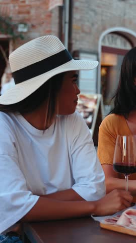 Vertical. Three cheerful female friends are laughing and enjoying a glass of red wine with a charcuterie board, spending quality time together in a picturesque Italian town