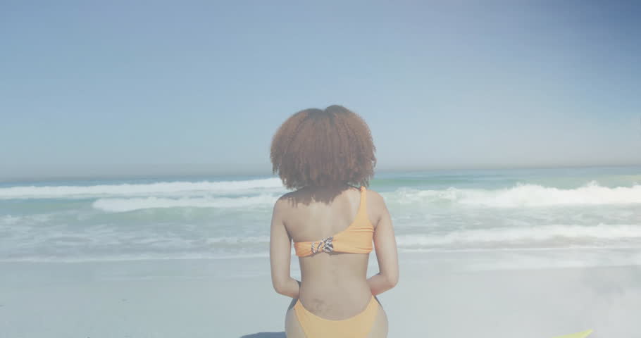 Woman shifting on yellow surfboard, leaning back and revealing fading flag overlay for marketing. Beach, ocean, coastal, leisure, resilience, patriotism, serenity