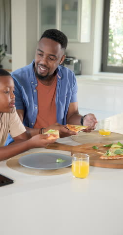 Vertical video: African American father and son eating pizza at counter reacting to basil, laughing. Family, leisure, rustic, candid, affectionate, culinary, vertical video