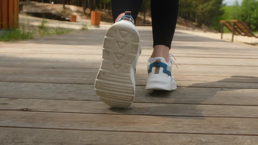 Close up woman feet running on wooden path in park, wearing modern white sneakers with blue details, highlight movement impact each stride. Woman running on wooden path in park wearing modern sneakers