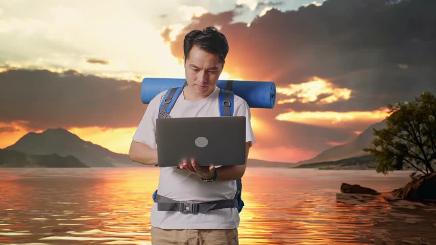 Asian Male Hiker With Mountaineering Backpack Using A Laptop At A Lake