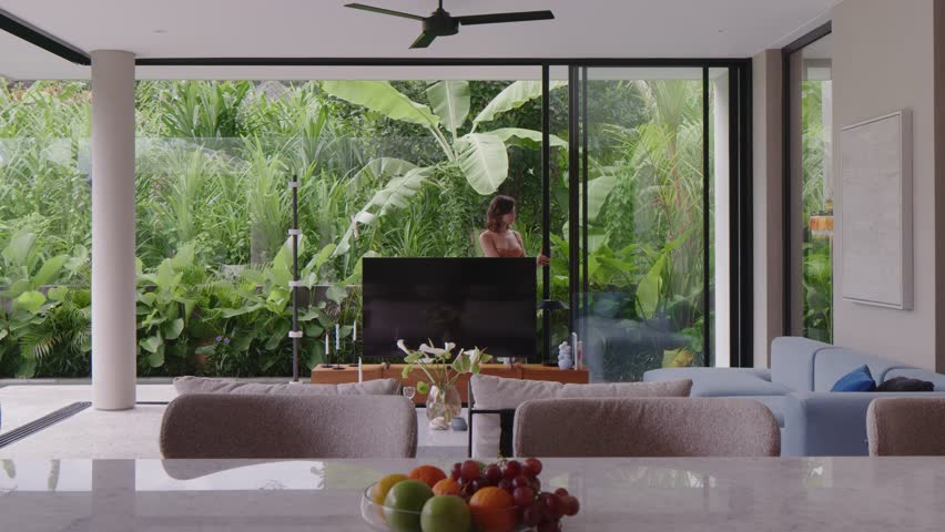 In a spacious stylish interior, a person enjoys the tranquility of lush greenery visible through large glass windows. Sunlight filters in, illuminating the peaceful atmosphere.
