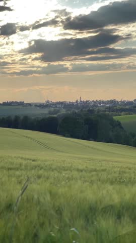 Rolling green fields lead towards a distant village with a prominent church steeple under a dramatic, cloudy sunset sky in rural Saxony, Germany.

