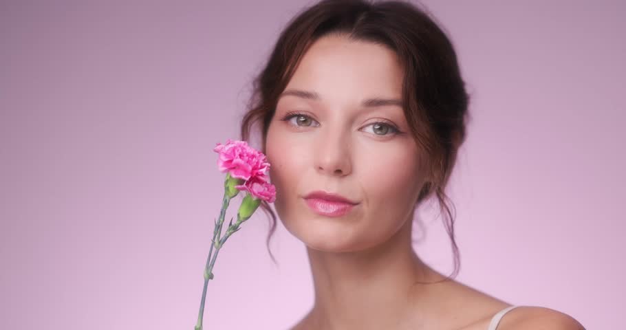 Beautiful woman gently touches her face with a pink flower, radiating softness and feminine beauty. Perfect for skincare, tenderness, fragrance, and wellness promo visuals.