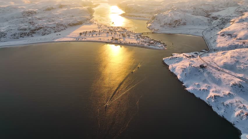 Aerial view of the Teriberka river and a snow-covered village reflecting the vibrant golden light of a winter sunset, Russia