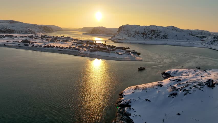 Aerial view of Teriberka village and river under a bright Arctic sunset, with a boat navigating the water, Russia