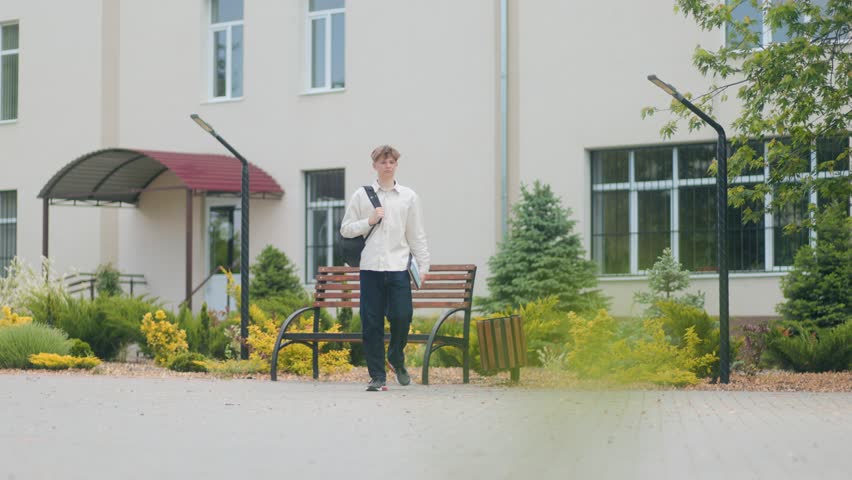Teenage boy walking alone through the schoolyard holding a book and backpack, captured full body in a natural setting outside the school during daytime