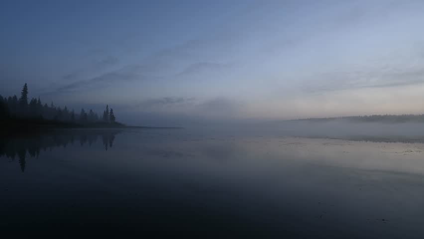 Timelapse of early morning mist blowing across a calm northern lake at blue hour with the water reflecting the trees and blue sky