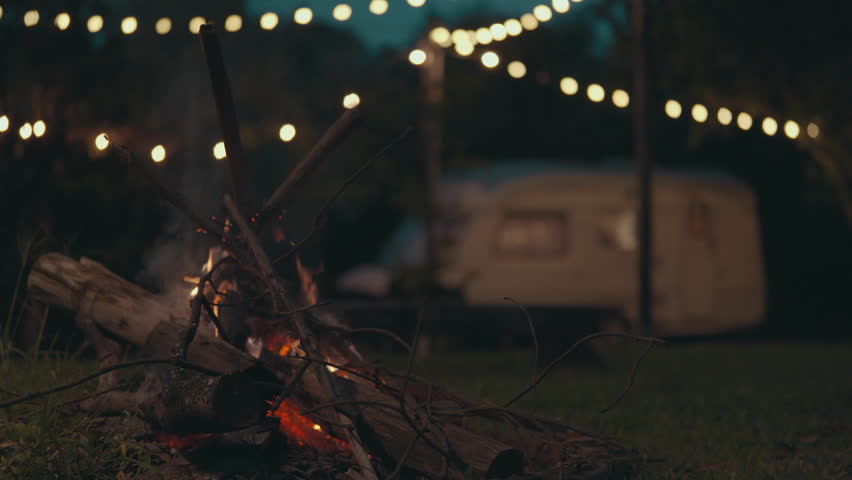 Camper is enjoying a bonfire near his cozy travel trailer in an autumn forest at night, Serene scene of a caravan with a campfire and string lights surrounding, set in a calming forest at dusk