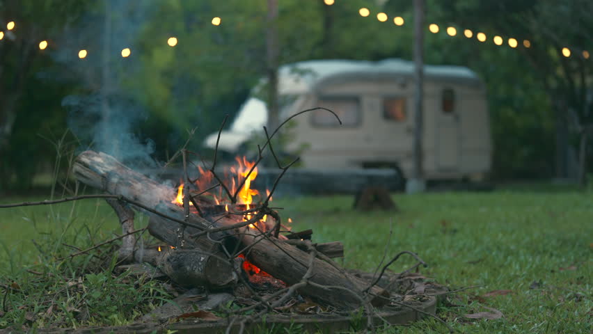 Camper is enjoying a bonfire near his cozy travel trailer in an autumn forest at night, Serene scene of a caravan with a campfire and string lights surrounding, set in a calming forest at dusk