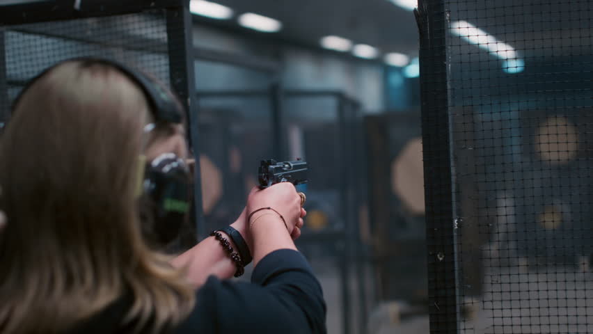 Woman with a gun fires a firearm at a shooting range. Female is engaged in sports shooting, wearing headphones. Pistol is a weapon. Cartridge case goes off in slow motion