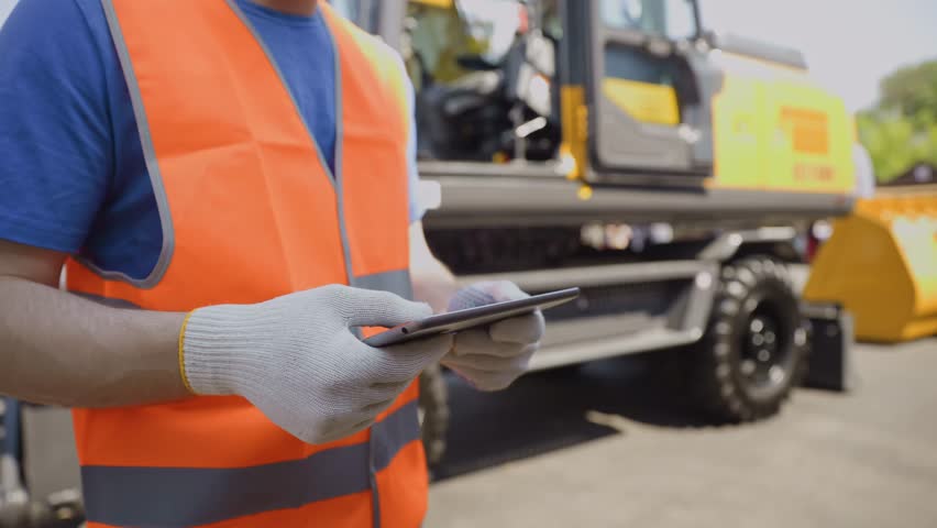 Industrial driver man engineer uses tablet to check online status of excavator at construction site.