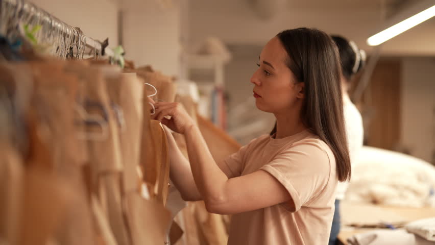Side view of young seamstress choosing sewing pattern hanging on clothes rack in bright atelier, preparing to create new clothing collection. Concept of garment manufacturing. Shooting in slow motion.