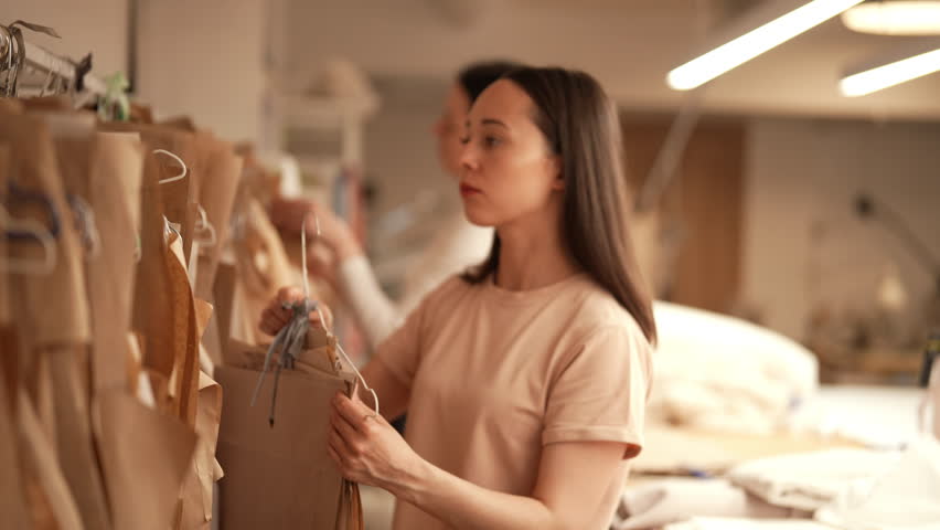 Side view of young seamstress choosing sewing pattern hanging on clothes rack in bright atelier, preparing to create new clothing collection. Concept of garment manufacturing. Shooting in slow motion.