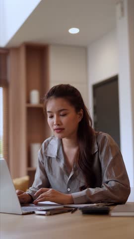 Beautiful Asian woman concentrates on personal finance management, using her laptop and calculator at the kitchen table in a cozy modern home setting.