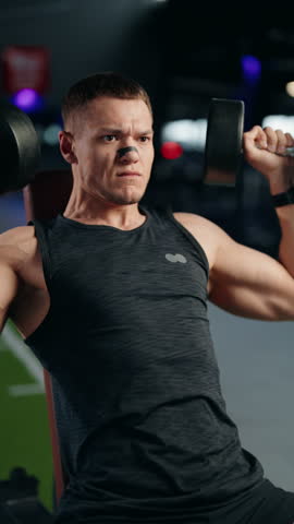 A muscular man performs dumbbell shoulder presses in a gym. He is focused and determined, pushing himself to the limit. This video captures the essence of fitness, strength, and dedication.