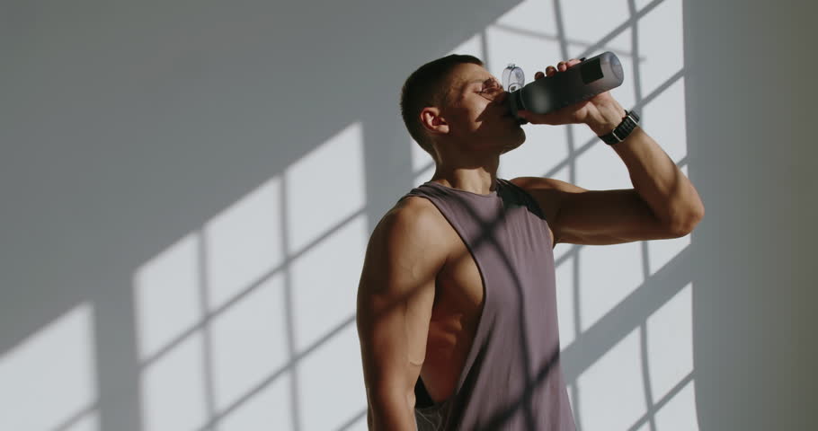 Athletic man hydrates while training in a bright studio, casting striking shadows. Young Athletic Man Drinking Water During Studio Workout