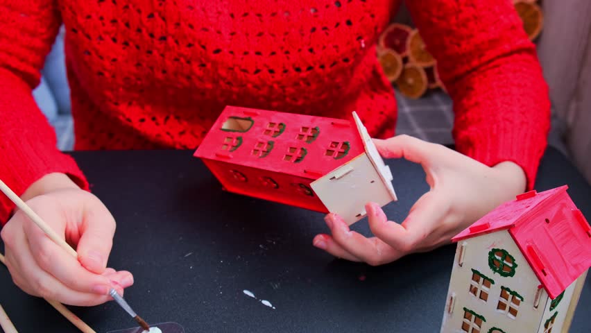 Unrecognizable woman in red sweater painting craft wooden houses for conscientious Merry Christmas decor. Sustainable celebration minimal responsible DIY holiday project. Step by step instruction