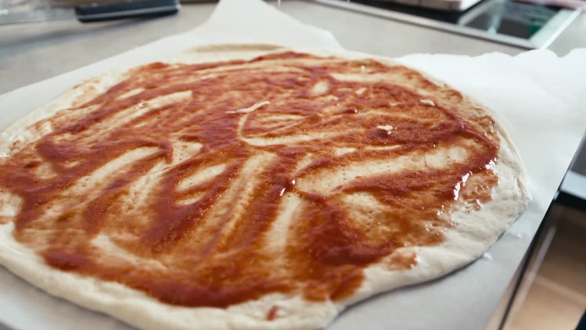 A person prepares a homemade pizza by spreading sauce on dough and evenly sprinkling cheese. This cozy kitchen scene showcases a love for cooking and crafting delicious meals.