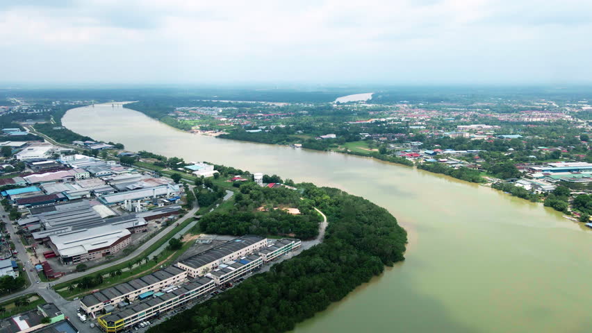 Aerial view of a wide river winding through an urban town with lush mangrove trees and industrial buildings on the riverbank. Peaceful water landscape from above.