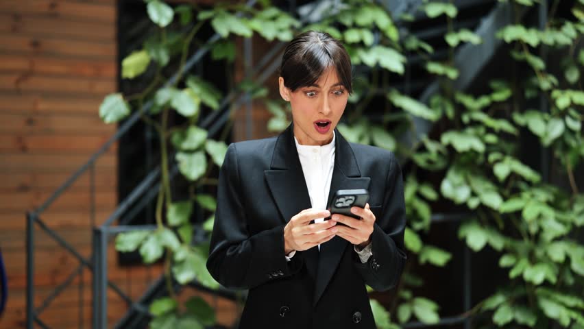 Excited businesswoman in suit rejoices at good news on phone. Stylish female feels happy at modern office. Lucky winner celebrates success in business.
