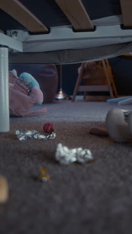 Vertical ground level shot of little African American girl looking for her toy on messy floor under bed using flashlight