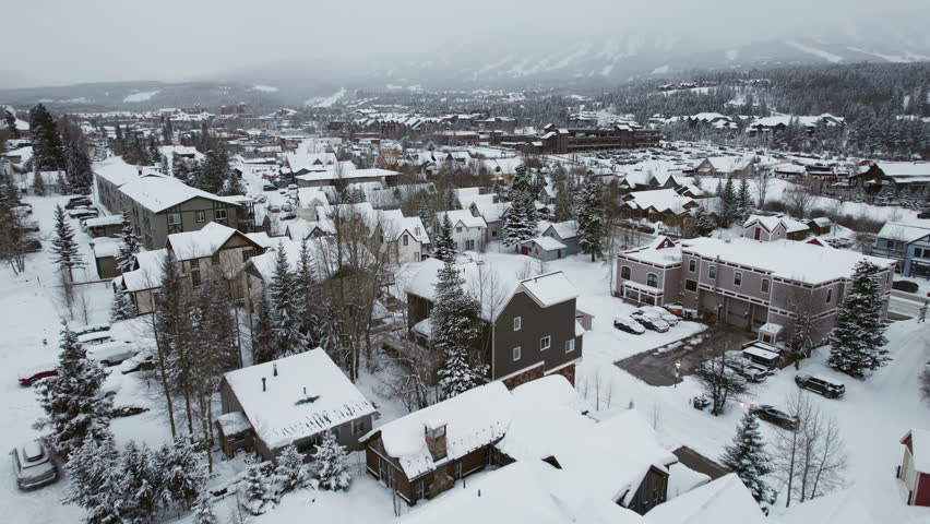 Dynamic aerial over winter cabins in Breckenridge, Colorado, past trees covered in snow, and a main road with cars carrying skis and snowboards into the popular mountain town and world famous resort.